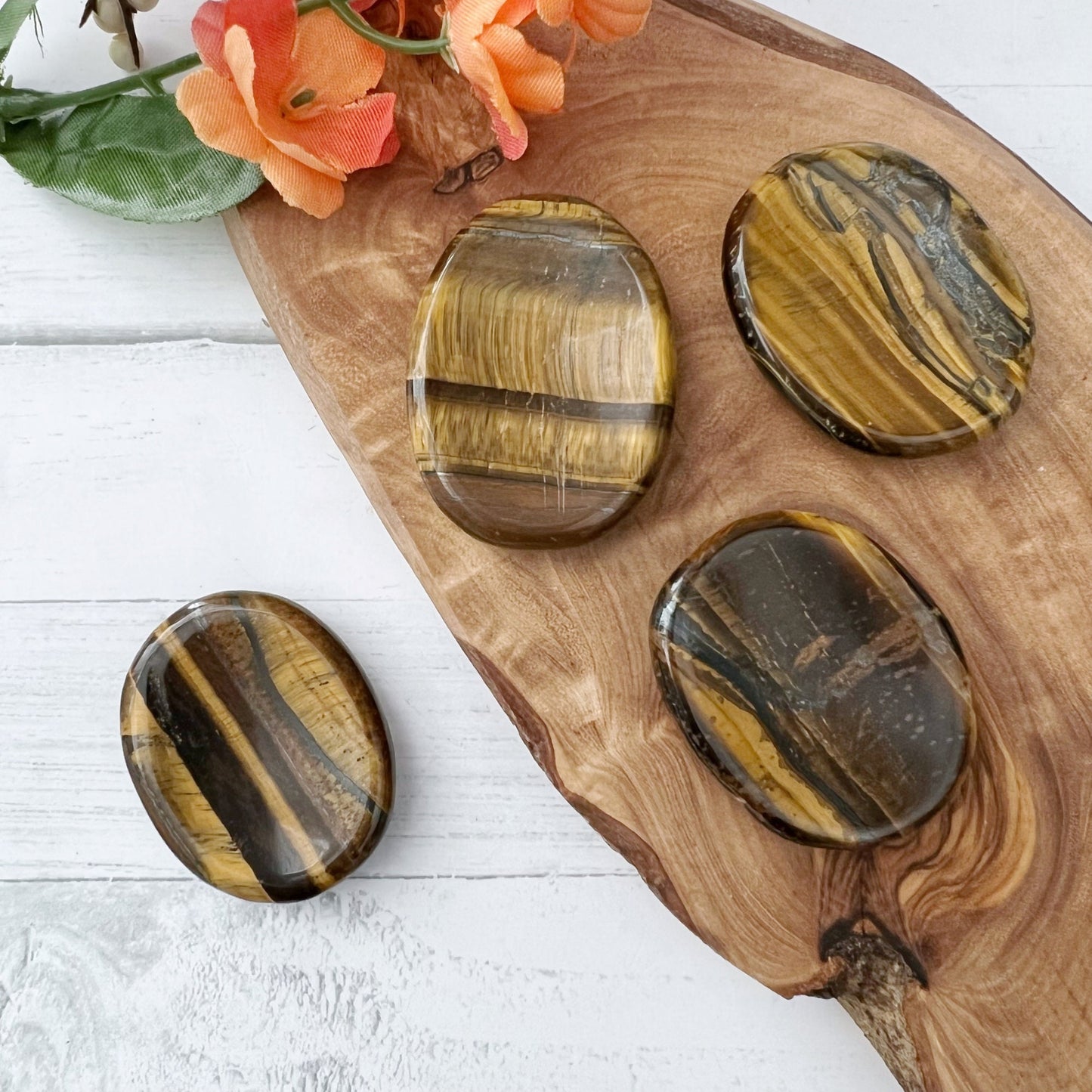 Four polished Tigers Eye Crystal Worry Stones with varying colors of gold, brown, and black are displayed on a wooden board. In the top left corner, an orange flower adds a touch of natural beauty to the arrangement on a light-colored surface, enhancing their appeal for crystal healing.