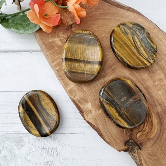 Four polished Tigers Eye Crystal Worry Stones with varying colors of gold, brown, and black are displayed on a wooden board. In the top left corner, an orange flower adds a touch of natural beauty to the arrangement on a light-colored surface, enhancing their appeal for crystal healing.