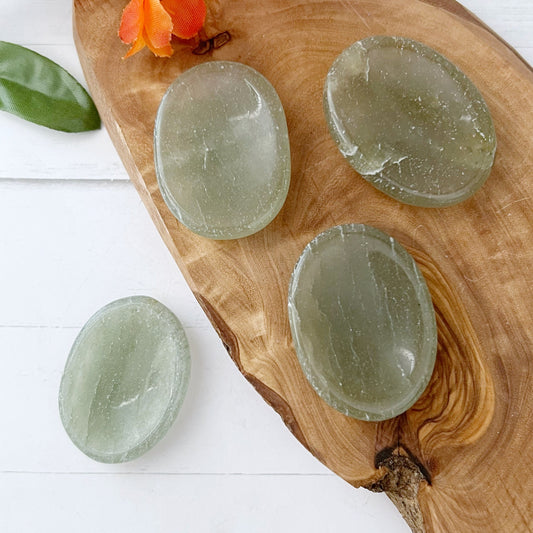 Four polished Green Aventurine Worry Stones are displayed on a wooden tray. One additional crystal rests off the tray on a white surface. A green leaf and an orange flower adorn the top left corner of the image, offering both natural beauty and stress relief for your senses.