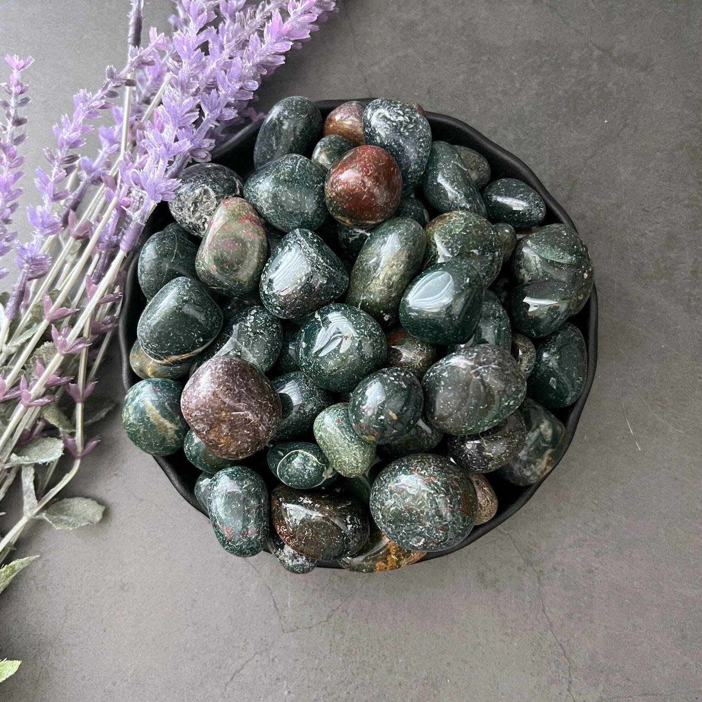 A black bowl filled with polished, multicolored stones, mostly dark green with some red and brown hues, sits on a dark gray surface. Sprigs of purple flowers lie next to the bowl, complementing the beauty of the Bloodstone Tumbled Stones known for enhancing intuition.