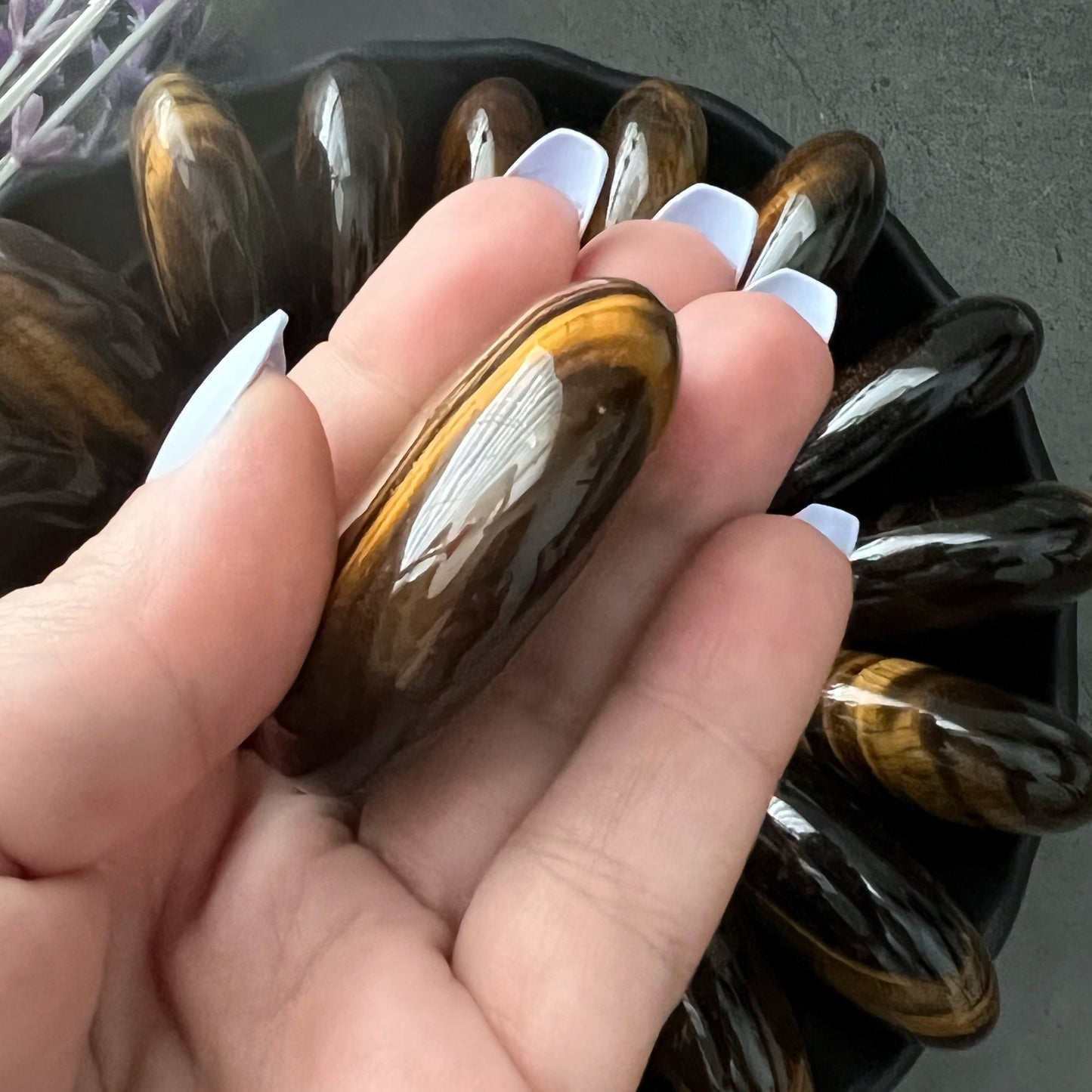 A black bowl filled with polished Tiger Eye Palm Stones arranged in a circular pattern creates a mesmerizing display. Two medium-sized, rounded Tiger Eye Palm Stones are at the center, surrounded by elongated oval-shaped stones. Lavender sprigs lie beside the bowl on the gray surface, adding a touch of serenity and wisdom.