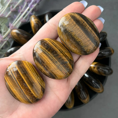 A black bowl filled with polished Tiger Eye Palm Stones arranged in a circular pattern creates a mesmerizing display. Two medium-sized, rounded Tiger Eye Palm Stones are at the center, surrounded by elongated oval-shaped stones. Lavender sprigs lie beside the bowl on the gray surface, adding a touch of serenity and wisdom.