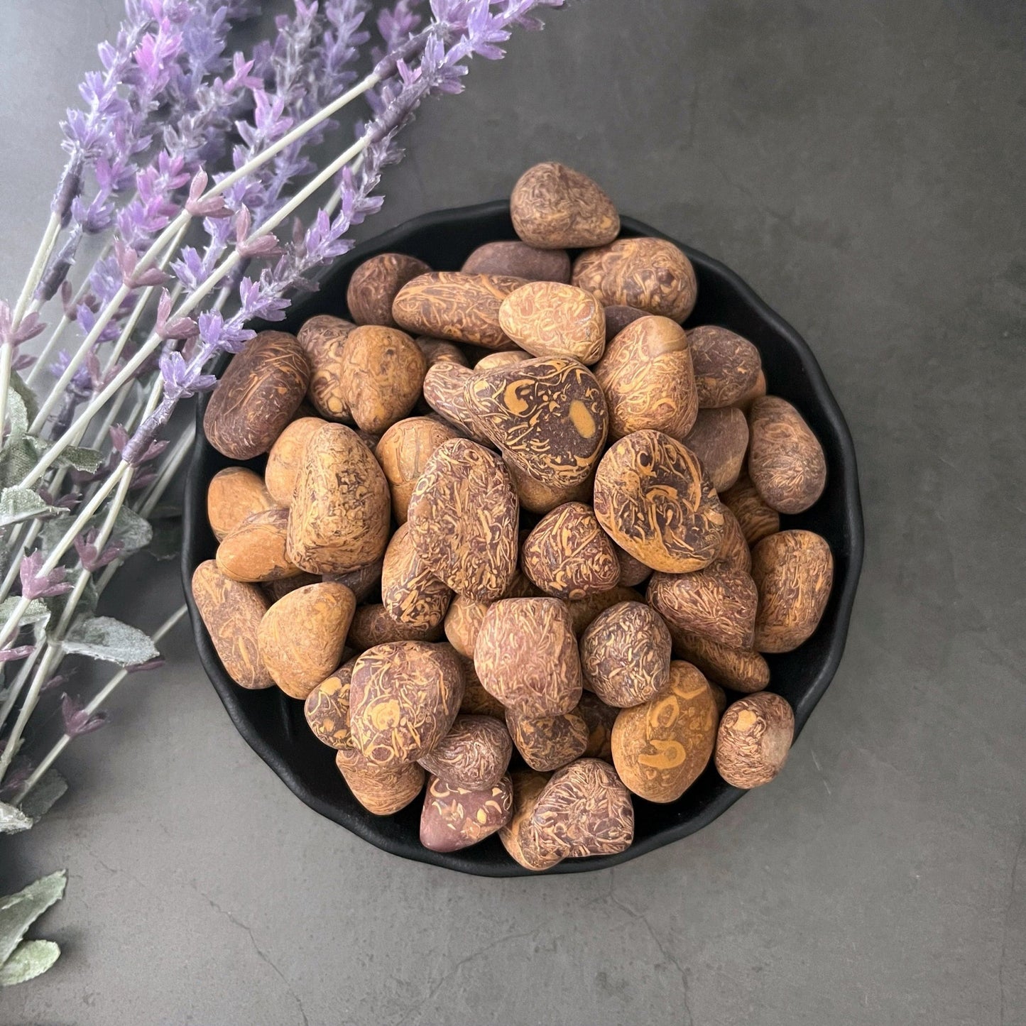 A black bowl filled with smooth, oval-shaped Calligraphy Jasper Tumbled Stones intricately etched with delicate patterns sits on a dark surface. To the left of the bowl, purple lavender sprigs lie diagonally, lending a touch of color to the serene composition.