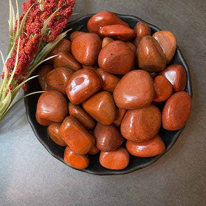 A black bowl filled with smooth, polished Red Jasper Tumbled Stones is placed on a gray surface. A sprig of red berries and greenish stems is laid beside the bowl, adding a touch of natural decor. This setup not only enhances the visual appeal but also resonates with Base Chakra and kundalini energy balancing.