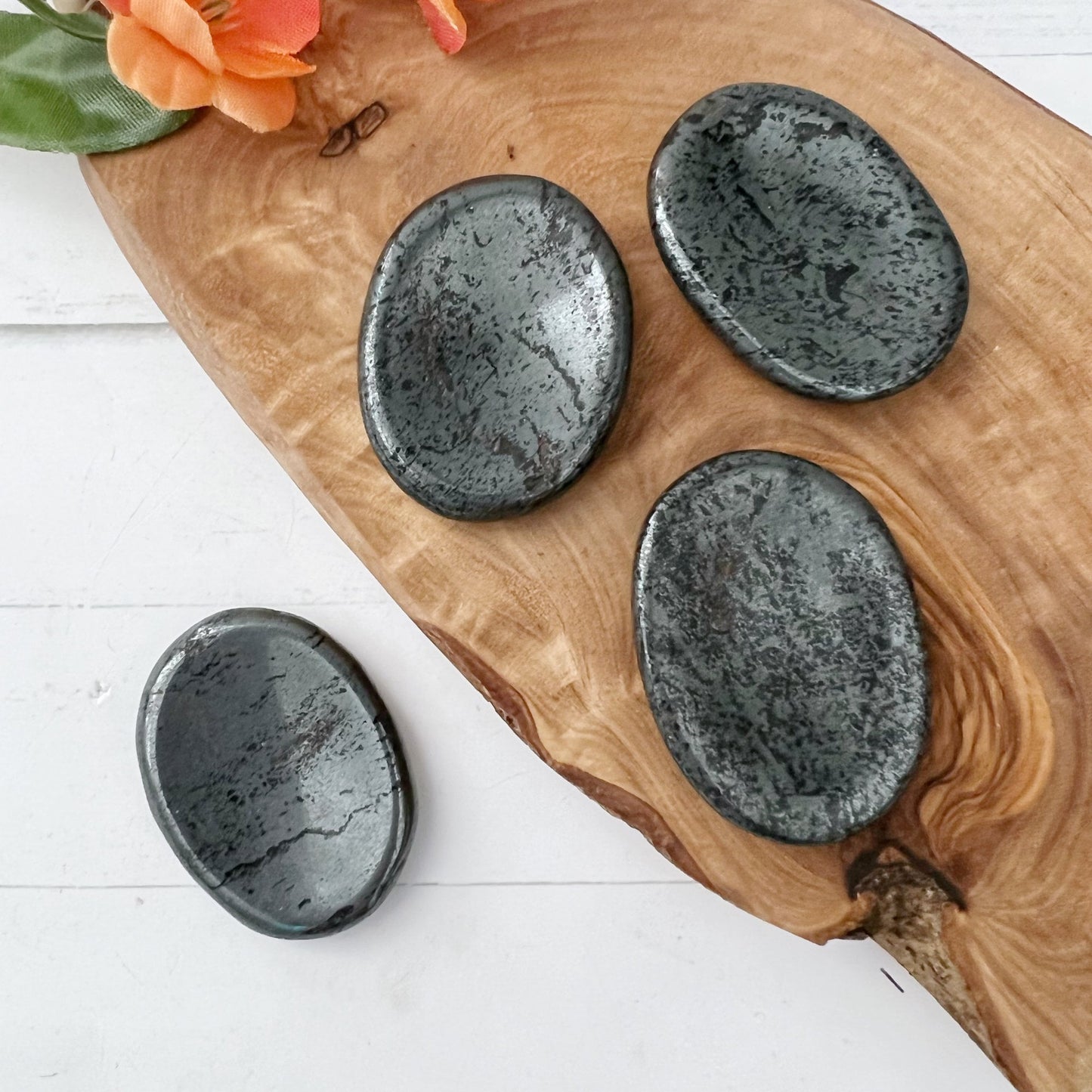 Four oval-shaped, polished Hematite Worry Stones are arranged on a wooden board. An orange flower is placed at the top left corner of this mindfulness accessory. The background is a white wooden surface, creating a serene and calming display.