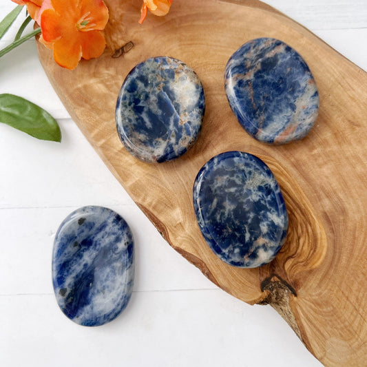 Four smooth blue and white stones, including a hand-polished Sodalite Worry Stone, are placed on a wooden board alongside vibrant orange flowers and green leaves. The stones have polished surfaces with varying patterns of deep blue and light white marbling. The scene is set on a white surface.