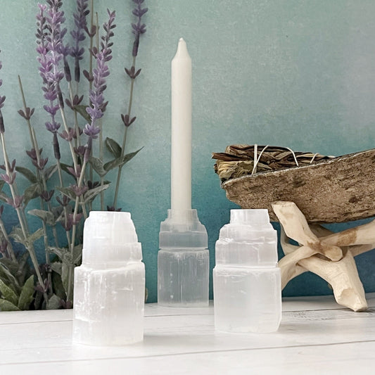 A serene setting featuring three selenite crystal towers in the foreground, a 2" Selenite Iceberg Mini Candle Holder for Ritual Chime Candles with a white candle standing upright, and dried lavender in the background. A small bundle of dried herbs rests on a wooden stand against a calming teal backdrop. Perfect for meditation work or candle rituals.