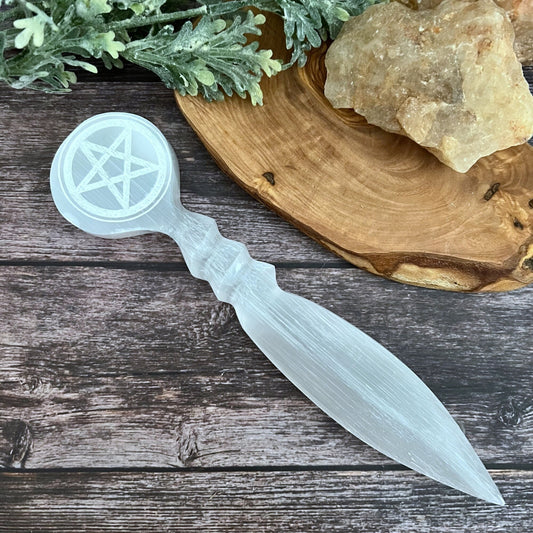 An ornate, clear, crystal-like 9.5" Selenite Pentacle Athame with a pentagram symbol engraved on the round handle lies on a wooden surface, accompanied by green foliage and rough, protective stones often used in Wiccan rituals.