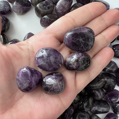 A black plate filled with polished Dark Dream Amethyst Tumbled Stones is placed on a dark surface. To the left of the plate, there are light pink rose flowers with green leaves. The amethyst stones, intuitively chosen for their stress-relieving properties, have a glossy finish and exhibit various shades of purple.