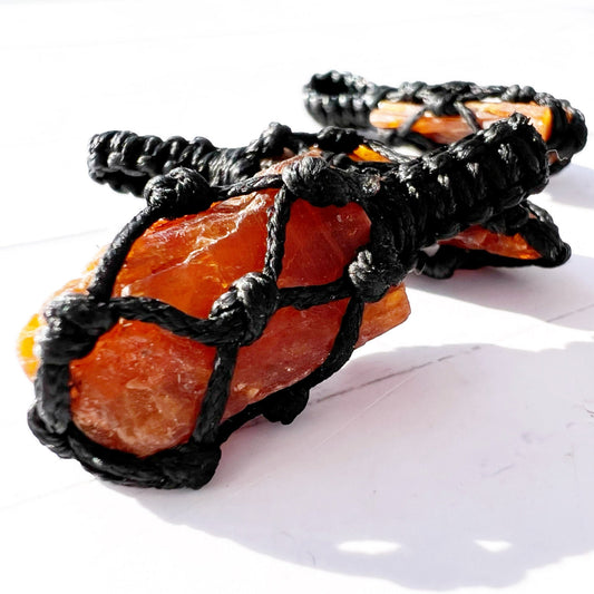 A Rough Orange Kyanite Crystal Thread Cage Pendant wrapped in an intricate black macrame netting is displayed against a white background. The detailed craftsmanship of the macrame cord securely encloses the irregularly shaped crystal, often associated with balancing the Sacral Chakra and enhancing creative thinking.