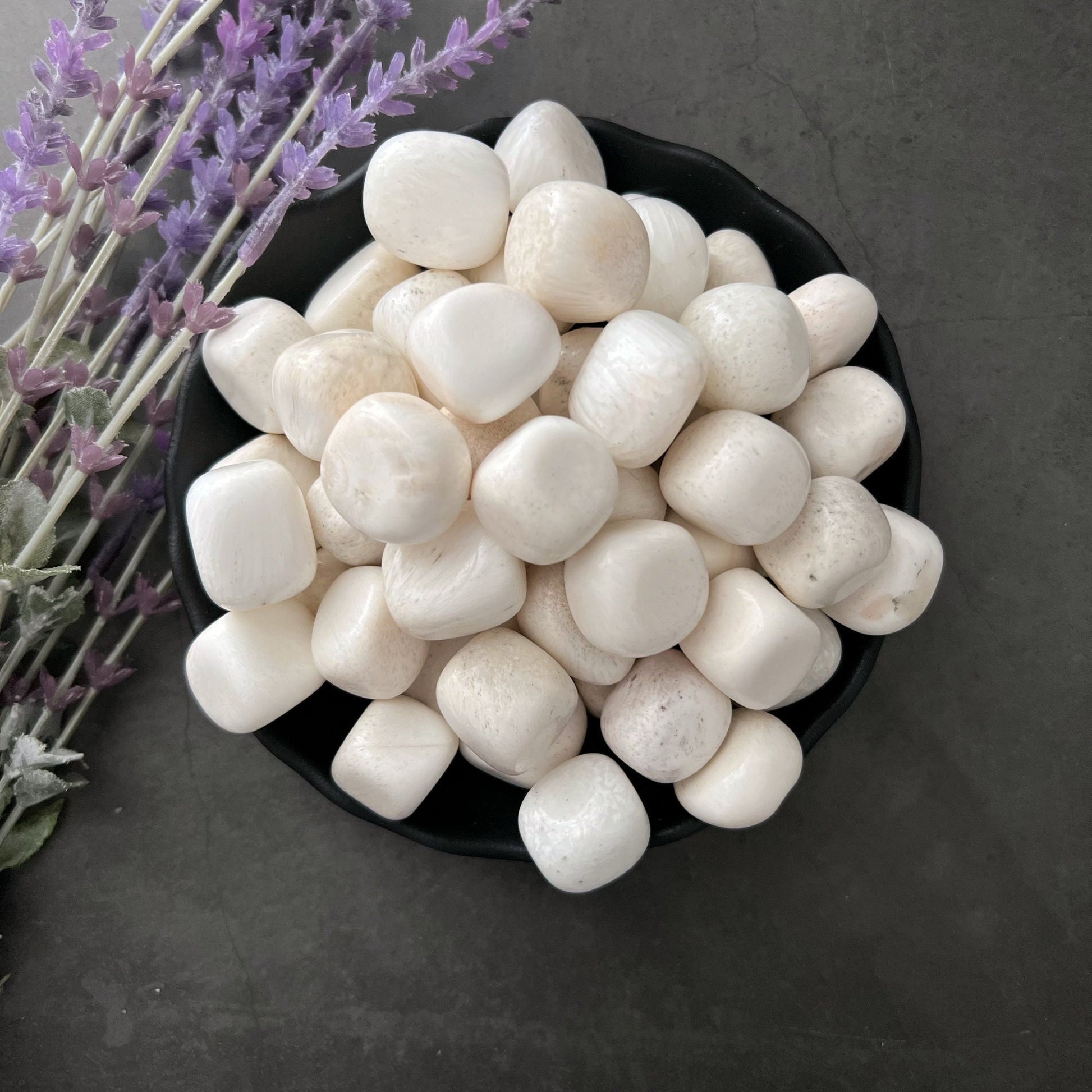 A black bowl filled with white, round, smooth pebbles and Scolecite Tumbled Stones is placed on a dark surface. To the left of the bowl, there is a bunch of lavender sprigs, adding a hint of color to the scene. This arrangement serves as an ideal meditation tool, promoting inner peace.