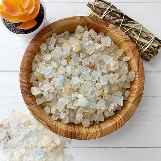 Close-up of a wooden bowl filled with various clear and light-colored crystal chips, including a few glimmers of Raw Natural Topaz Chips, on a white wooden surface. To the left, some crystals are scattered outside the bowl. In the background, there's a bundle of tied sticks and a small potted succulent.