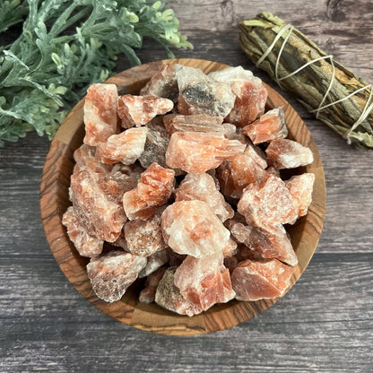 A wooden bowl filled with rough, pink-orange Raw Red Calcite Stones rests on a wooden surface. To the right of the bowl is a bundle of sage wrapped in twine, while to the left lies a small bunch of green foliage, all harmonizing to balance your root chakra and enhance increased energy.