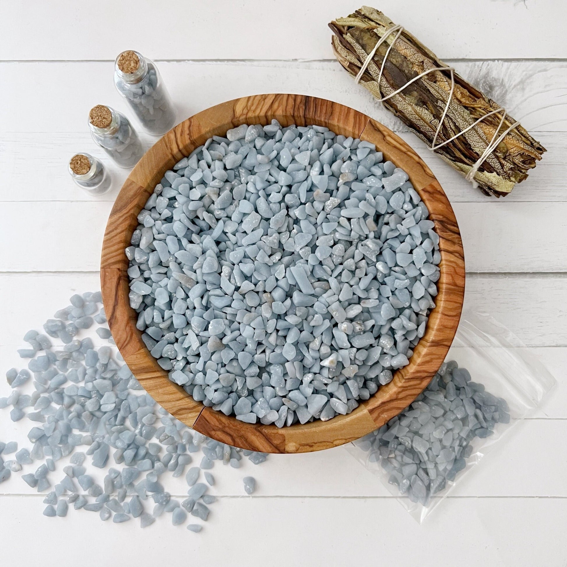 A wooden bowl filled with small, light blue Angelite Crystal Chips sits on a white wooden surface. Some stones are scattered around the bowl, three small glass bottles filled with stones are on the left, and a clear plastic bag containing stones is on the bottom right. A bundle of sage for spiritual healing is tied with string on the top right.