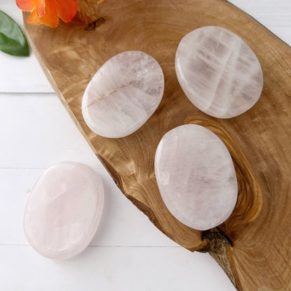 Four polished Rose Quartz Worry Stones rest on a wooden board. The board features swirling wood grain patterns, and a small orange flower with a green leaf is partially visible at the edge. The light-colored background enhances the soft pink hue of these stones, known for their emotional healing properties.