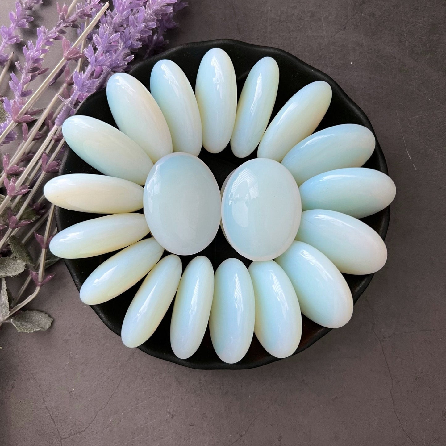 A black dish is arranged with smooth, oval-shaped 1.5" Opalite Palm Stones, radiating like petals around a central pair. Known for their healing properties, these milky white gemstones sit gracefully beside lavender sprigs on a gray surface.