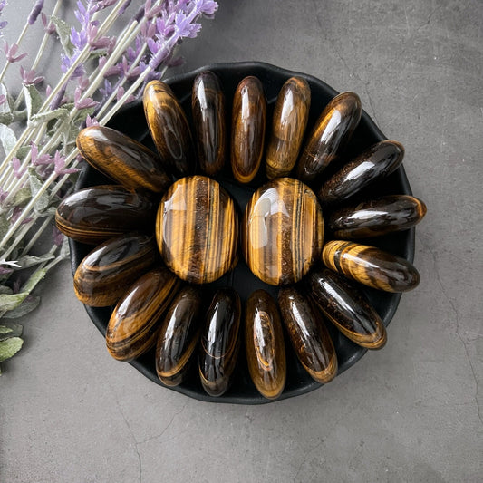 A black bowl filled with polished Tiger Eye Palm Stones arranged in a circular pattern creates a mesmerizing display. Two medium-sized, rounded Tiger Eye Palm Stones are at the center, surrounded by elongated oval-shaped stones. Lavender sprigs lie beside the bowl on the gray surface, adding a touch of serenity and wisdom.