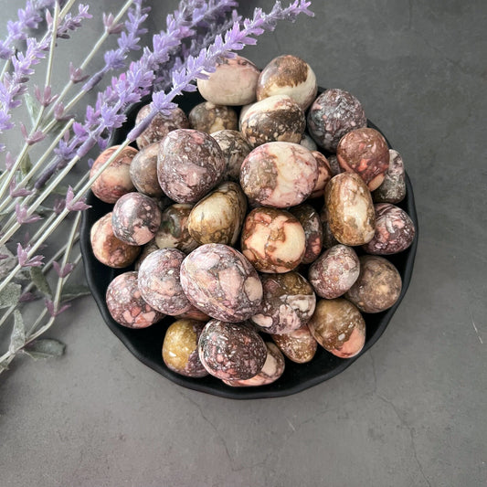 A black bowl filled with polished, multicolored stones featuring tones of purple, brown, and cream exudes calming energy. Among the stones are Pink Porcelain Jasper Tumbled Stones. The bowl sits on a dark surface next to sprigs of purple lavender flowers.