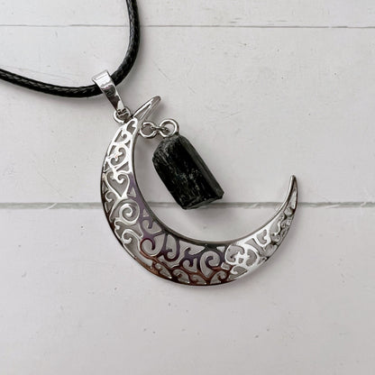 A Silver Crescent Moon & Black Tourmaline Pendant with intricate filigree design hangs from a black cord necklace, featuring a rough Black Tourmaline pendant for its protective properties. The background is a light, textured surface.