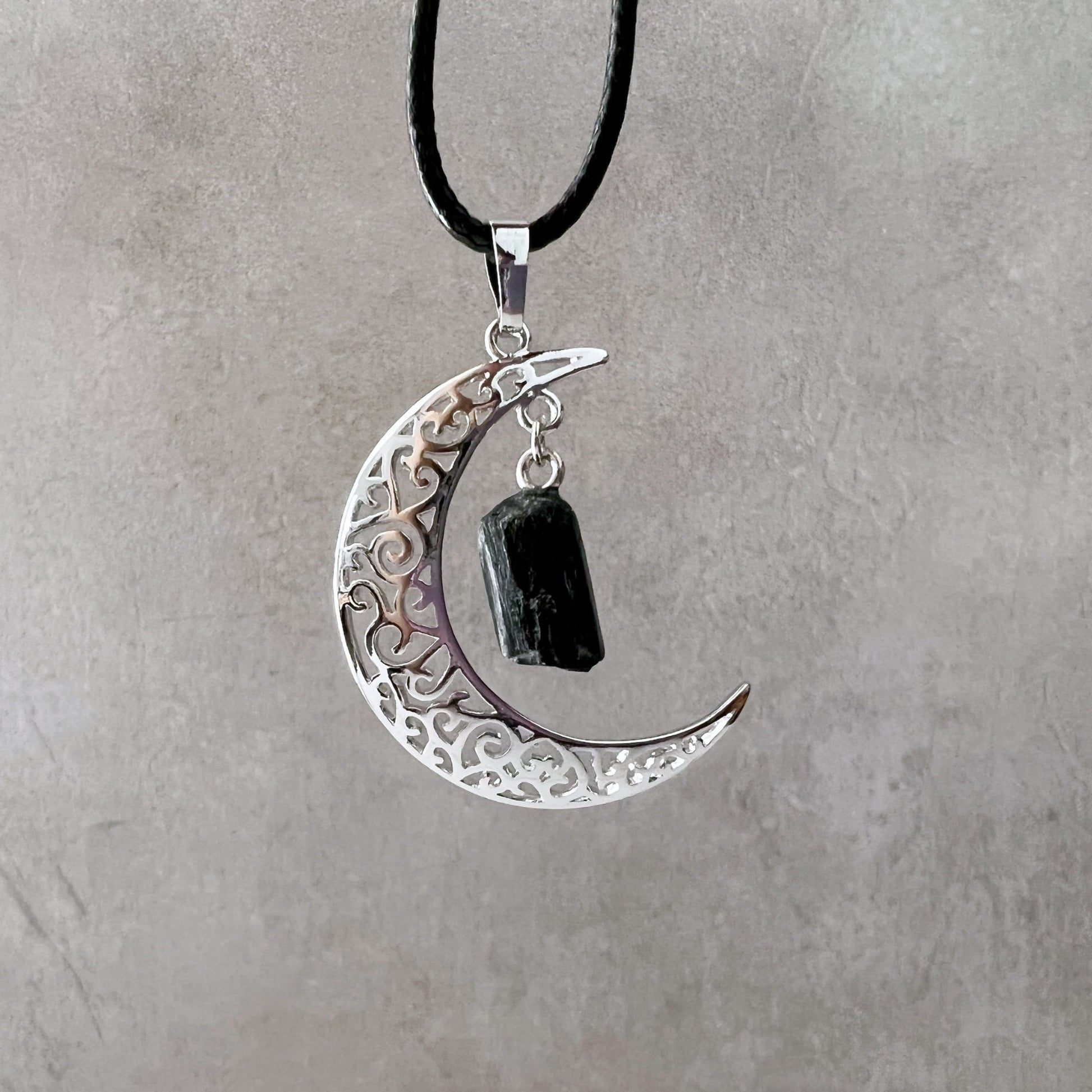 A Silver Crescent Moon & Black Tourmaline Pendant with intricate filigree design hangs from a black cord necklace, featuring a rough Black Tourmaline pendant for its protective properties. The background is a light, textured surface.