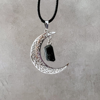 A Silver Crescent Moon & Black Tourmaline Pendant with intricate filigree design hangs from a black cord necklace, featuring a rough Black Tourmaline pendant for its protective properties. The background is a light, textured surface.