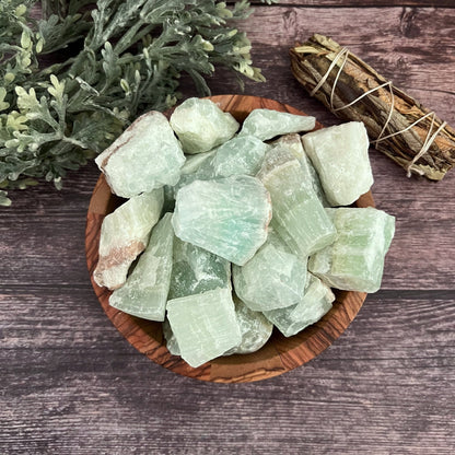 A wooden bowl filled with large, rough Raw Pistachio Green Calcite Stones sits on a wooden surface. A bundle of sage is tied with twine and placed next to the bowl, symbolizing abundance and prosperity. Green foliage partially frames the top left corner of the scene, adding a touch of natural elegance.