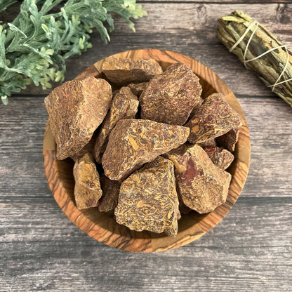 A wooden bowl containing chunks of Raw Calligraphy Jasper Stones, a striking reddish-brown meditation crystal, is placed on a weathered wooden surface. To the left, there is greenery, and to the right, there is a bundle of dried sage wrapped in twine.