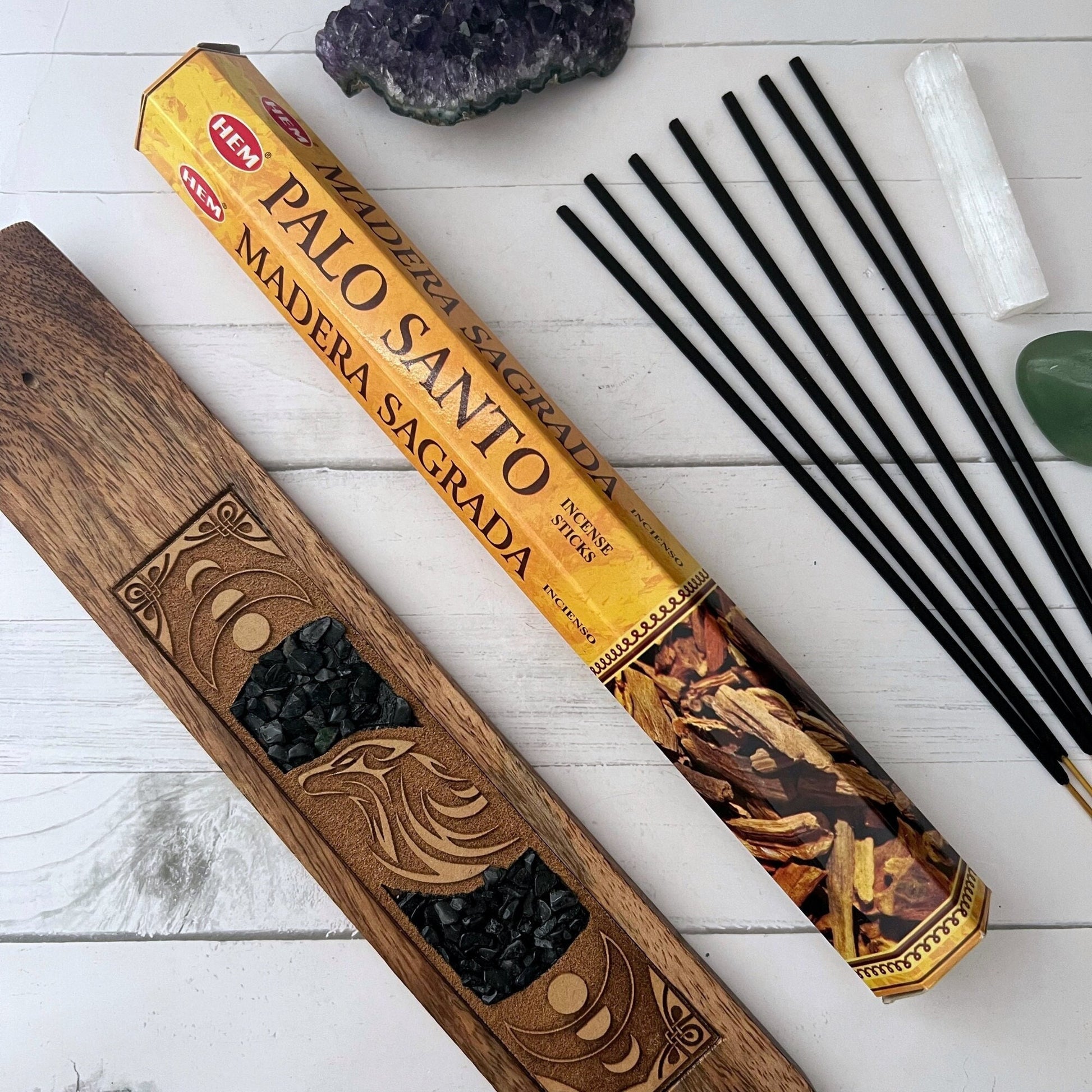 A standing rectangular box and a round container both labeled "Palo Santo Incense Sticks." The box displays an image of palo santo wood sticks, ideal for meditation, and text indicating the product is handcrafted in India. The round container is similarly branded and colored, perfect for enhancing your practice.