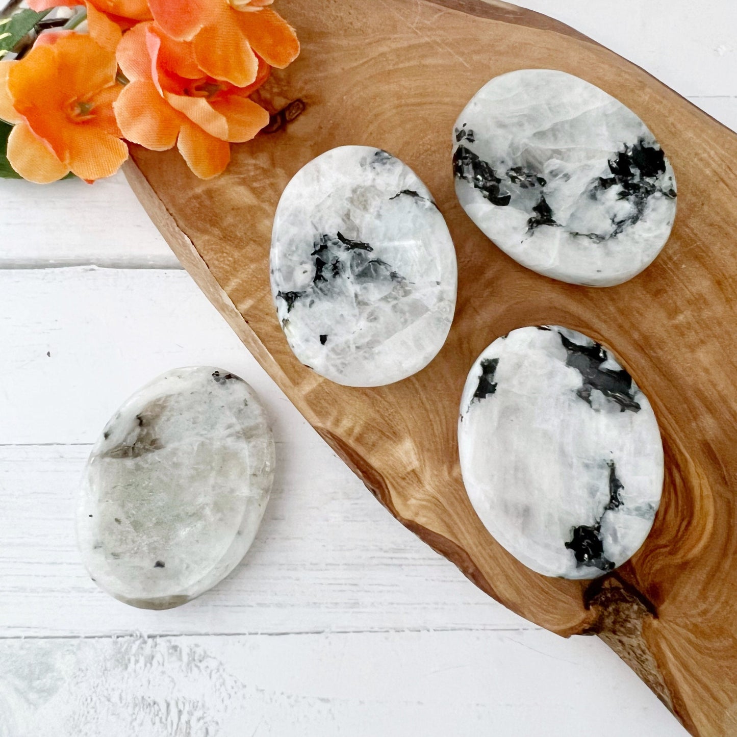 Four polished white Rainbow Moonstone Worry Stones with black and gray marbling lie on a wooden tray. An orange flower decorates the tray's top left corner. The tray and stones are on a white wooden surface, evoking thoughts of psychic protection and tranquility.