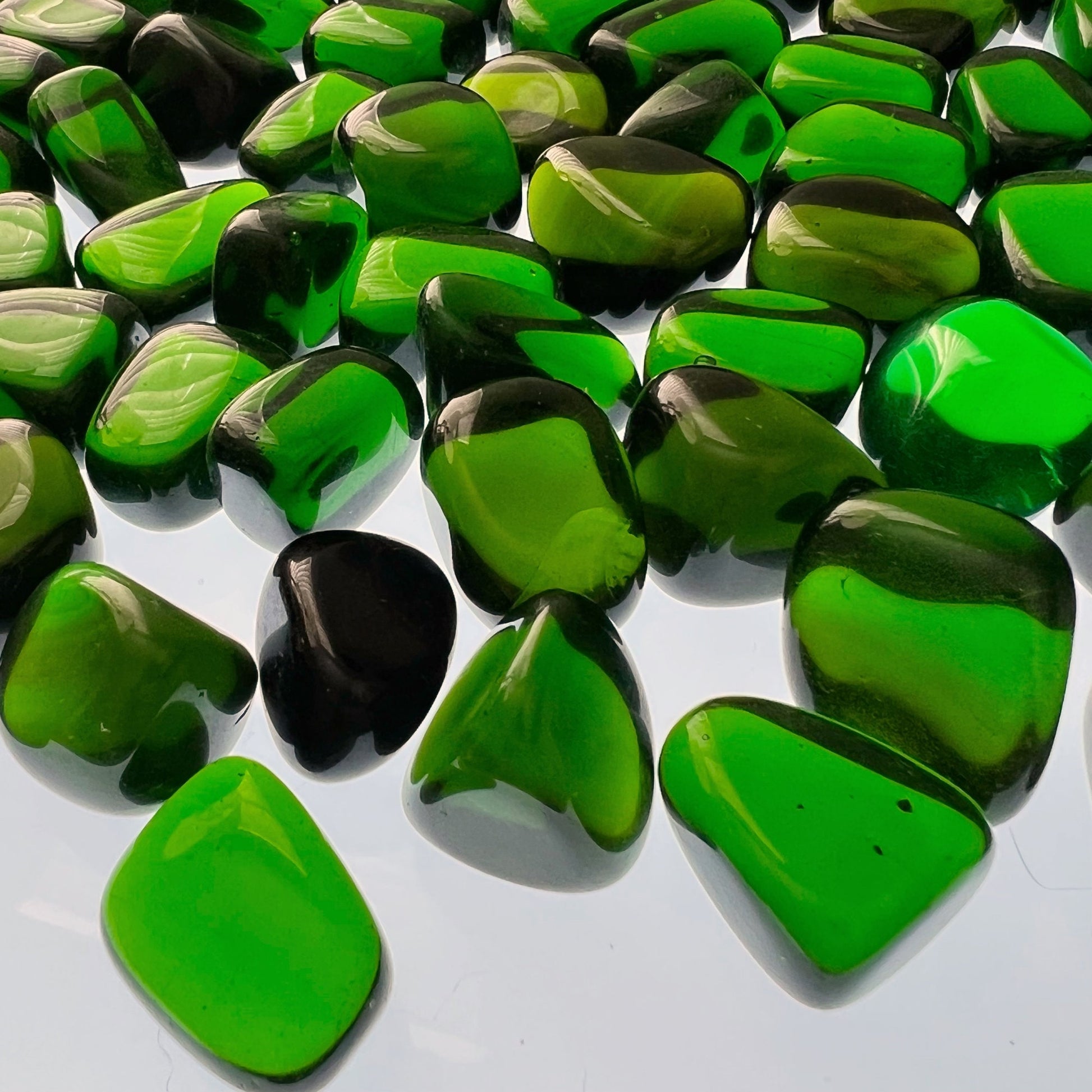 A black bowl filled with polished Green Obsidian Tumbled Stones with occasional green highlights. The bowl is positioned on a gray surface, and there are sprigs of purple flowers lying next to it. The scene, perfect for Reiki healing and heart chakra alignment, is elegantly arranged with natural elements.