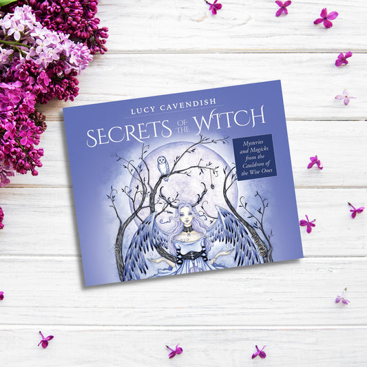 A product titled "Secrets of the Witch Oracle Cards: Mysteries and Magicks from the Cauldron of the Wise One" by Lucy Cavendish lies on a white wooden surface. The cover features a mystical illustration of a witch with flowing hair and an owl perched on a tree branch. Flowers, leaves, and small purple petals are scattered around the product, evoking the essence of her divination deck.