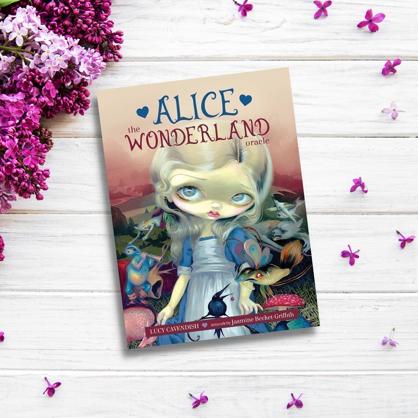 A book titled "Alice: The Wonderland Oracle Cards by Lucy Cavendish," illustrated by Jasmine Becket-Griffith, is placed on a white wooden surface, surrounded by purple flowers. The cover features a doll-like girl with blond hair and blue eyes, dressed in blue and white, standing with various fantasy creatures in a whimsical scene.