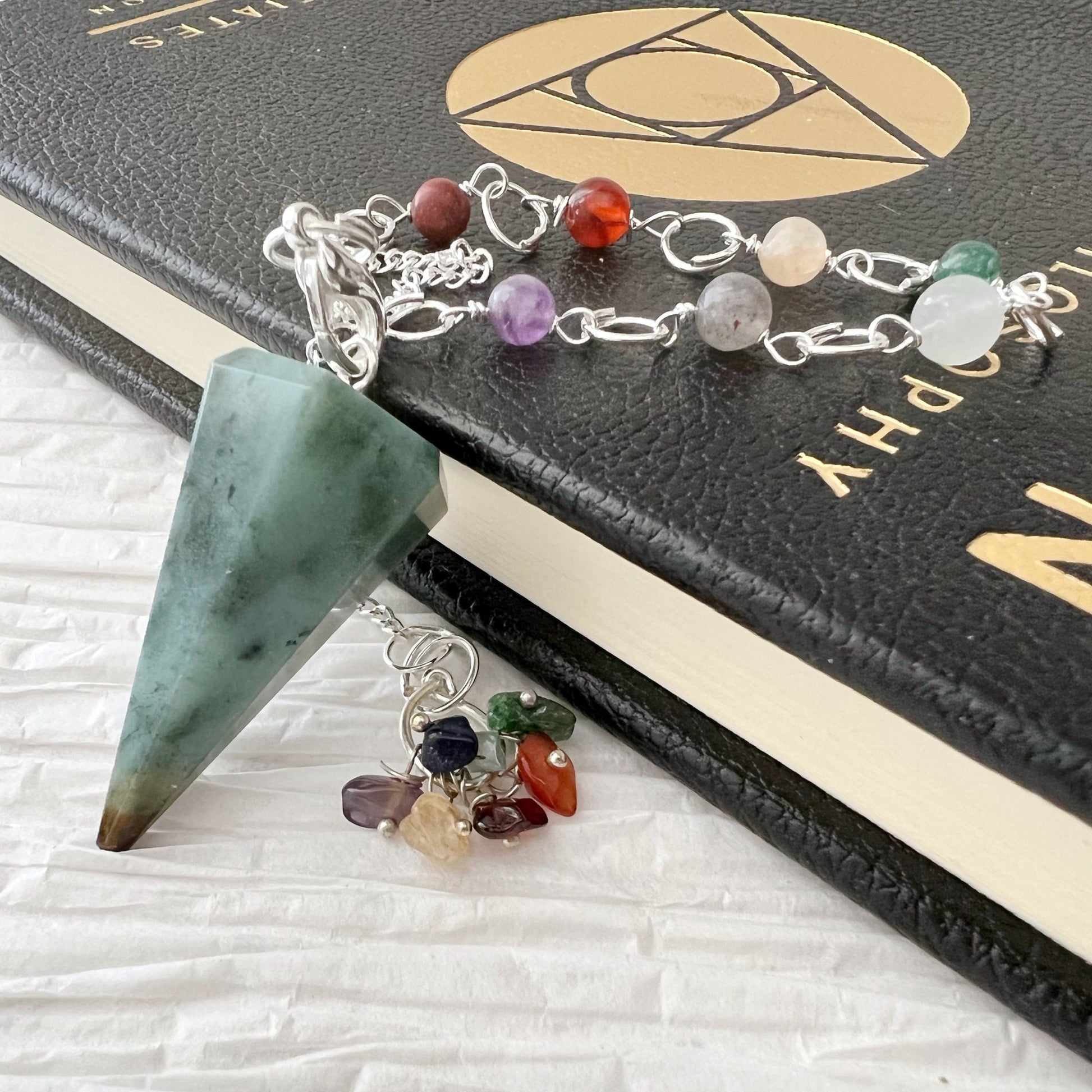 A Green Opal Hexagonal Pendulum attached to a bracelet with assorted gemstones rests on a black 'Numerology' book. The bracelet features stones of various colors such as red, purple, and clear and is placed on a white textured surface.