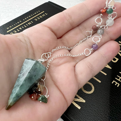 A Green Opal Hexagonal Pendulum attached to a bracelet with assorted gemstones rests on a black 'Numerology' book. The bracelet features stones of various colors such as red, purple, and clear and is placed on a white textured surface.