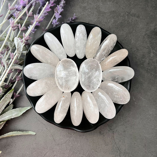 A black dish holds an arrangement of smooth, elongated Clear Quartz Palm Stones and rose quartz palm stones, forming a petal-like pattern. The dish is placed on a gray surface, alongside sprigs of lavender and sage leaves, emphasizing a serene ambiance ideal for healing.