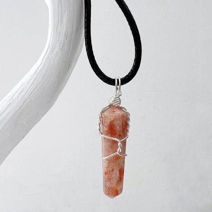 A 1.5" Double Terminated Sunstone Point Pendant consisting of an orange and white crystal wrapped in silver wire hangs from a black cord, evoking self-empowerment jewelry. The background is a plain white surface.