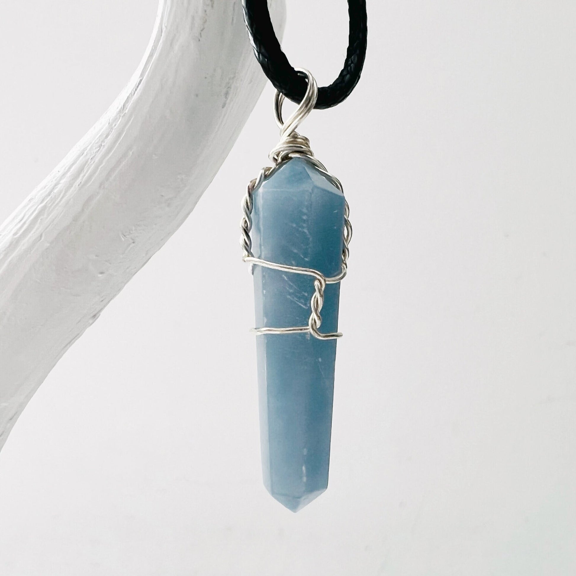 A 1.5" Double Terminated Angelite Point Pendant, wrapped with silver wire, hangs from a black cord. The pendant, known for aiding psychic healing and telepathic communication, is displayed against a plain white background with part of a white branch visible on the left side.