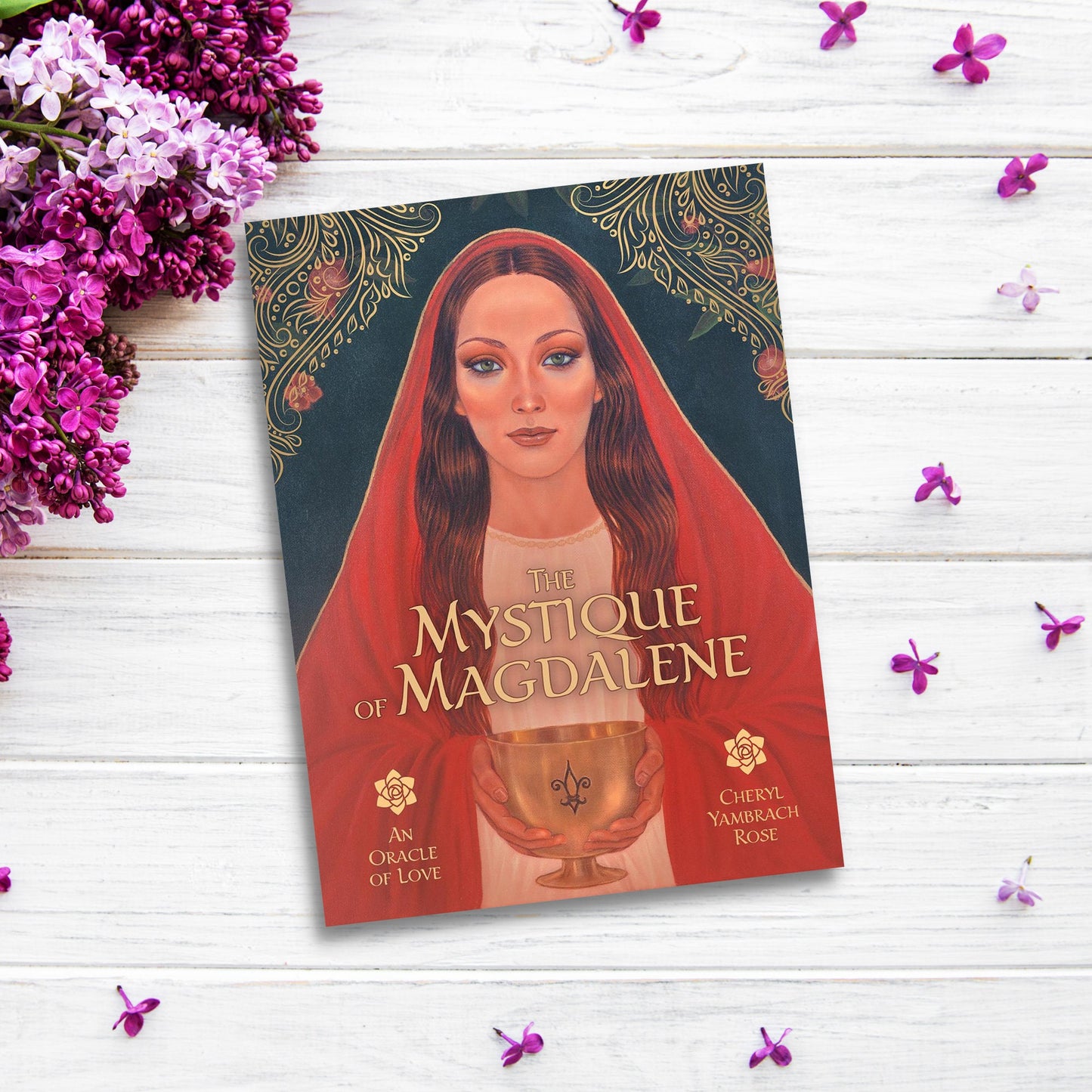 A book titled "Mystique of Magdalene" by Cheryl Yambrach Rose is displayed against a white wooden background. The cover features an illustrated woman in a red cloak holding a golden cup, with purple flowers arranged around the book, evoking the essence of Mary Magdalene.