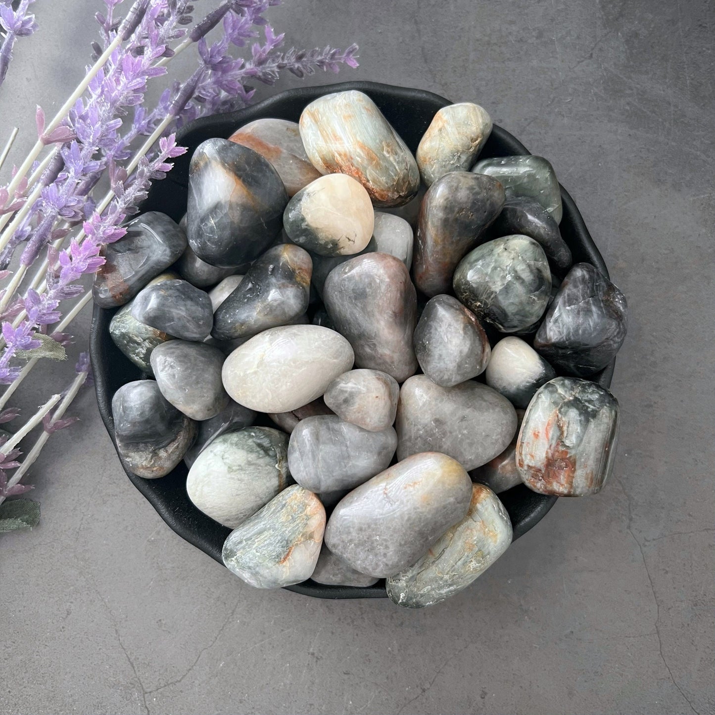 A black bowl filled with polished stones in various shades of gray, white, and subtle touches of green and brown. Among them lies a Cat's Eye Quartz Tumbled Stone, known for its metaphysical powers. To the left of the bowl, there is a sprig of lavender. The background is a smooth, gray surface.