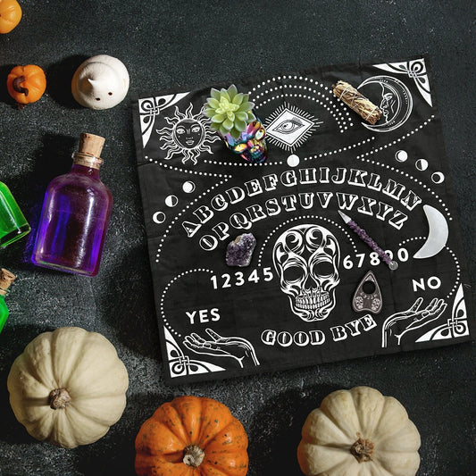 An overhead view of a mystical scene featuring a black Spirit Altar Cloth adorned with white lettering and symbols, such as the sun, moon, and an eye. The cloth is surrounded by small pumpkins, colorful potion bottles, crystals, an altar cloth, and a divination planchette.