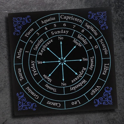 A black astrology board, akin to a 12" x 12" Embroided Velvet Zodiac Pendulum Mat, rests on the table, surrounded by small pumpkins and colorful potion bottles. This divination tool features a zodiac wheel with responses like "Yes," "No," and "Maybe," along with days of the week and calendar months.