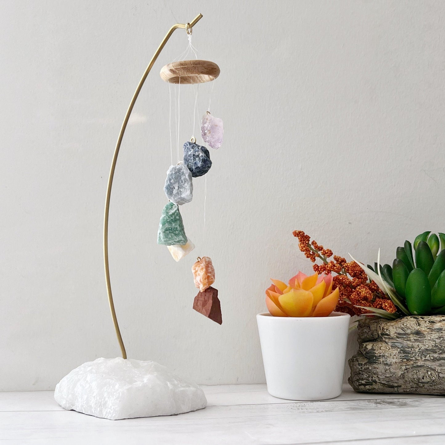 A decorative hanging display features a 7 Chakra Crystal Table Chime suspended on strings from a wooden ring, held by a curved metal stand anchored in a white stone base. Nearby are two potted succulent plants, one in a white pot and the other in a wooden container.