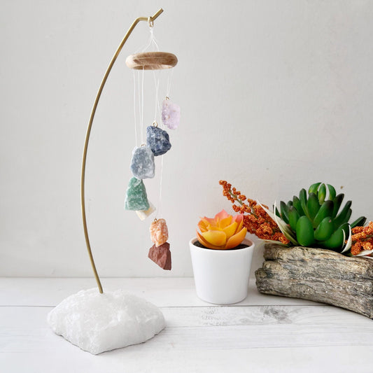 A decorative hanging display features a 7 Chakra Crystal Table Chime suspended on strings from a wooden ring, held by a curved metal stand anchored in a white stone base. Nearby are two potted succulent plants, one in a white pot and the other in a wooden container.