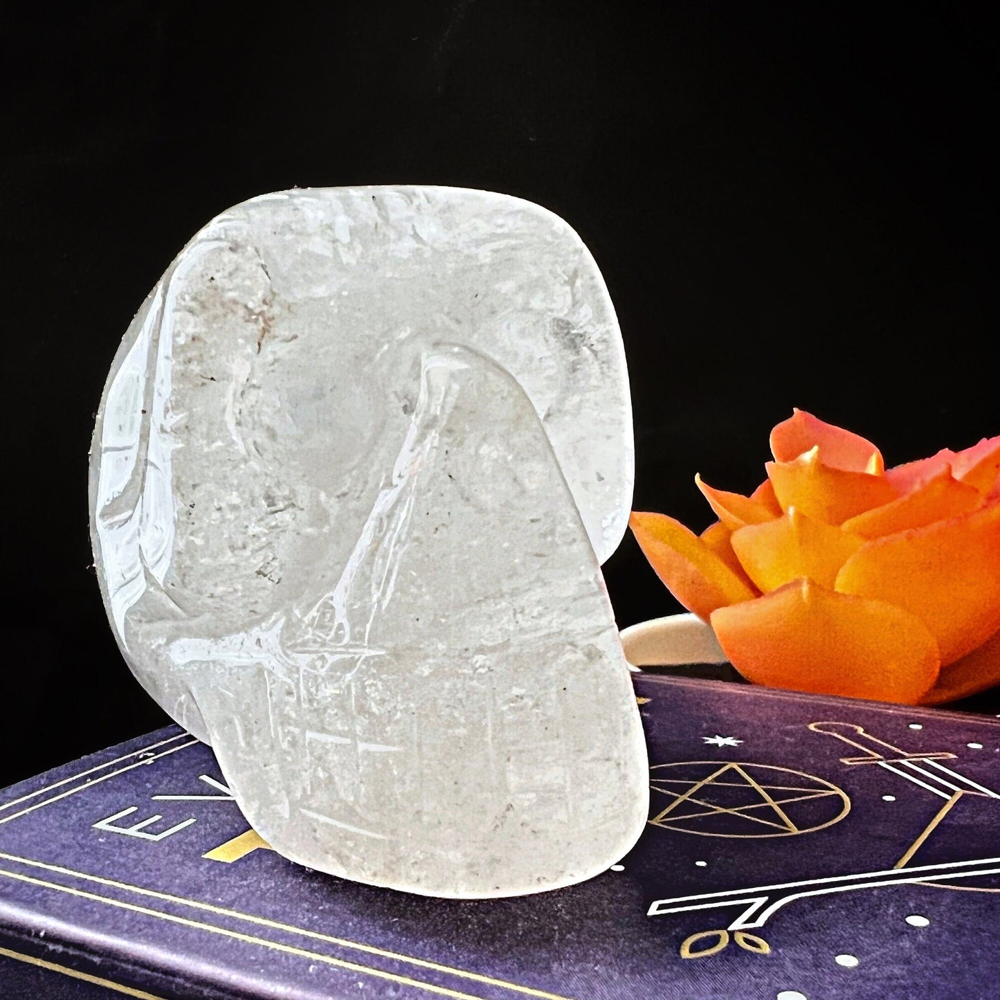 A smooth, translucent 2" Clear Quartz Crystal Skull sits on a purple book adorned with golden astrological symbols. An orange flower is placed beside the sculpture, providing a vivid contrast against the dark background, infusing the scene with serene Reiki energy.