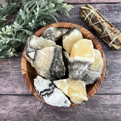 A wooden bowl contains large, rough chunks of Raw Zebra Calcite Stones in various shades of white, beige, and grey. The bowl is placed on a wooden surface, with a bundle of sage and a green leafy plant nearby, inviting transformational energies into the space.