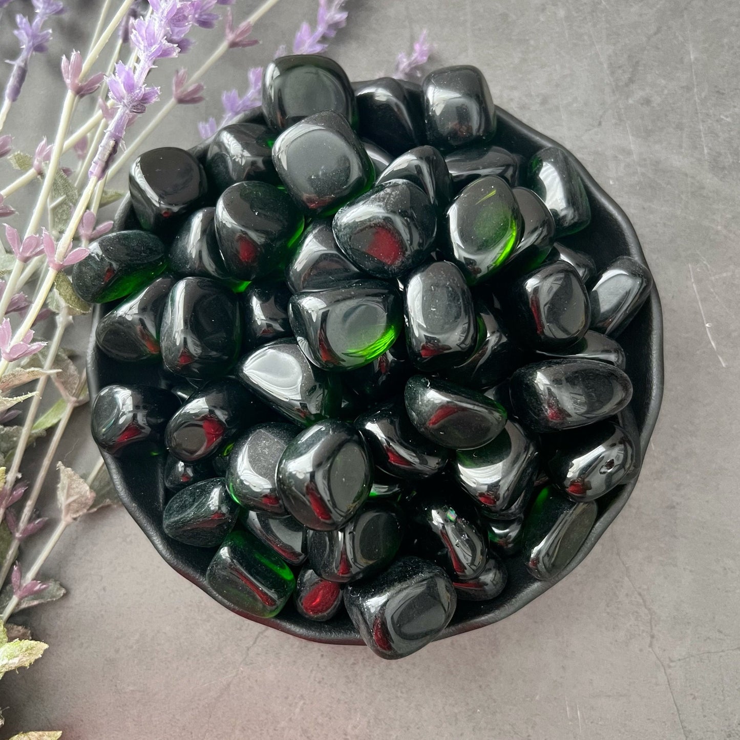 A black bowl filled with polished Green Obsidian Tumbled Stones with occasional green highlights. The bowl is positioned on a gray surface, and there are sprigs of purple flowers lying next to it. The scene, perfect for Reiki healing and heart chakra alignment, is elegantly arranged with natural elements.