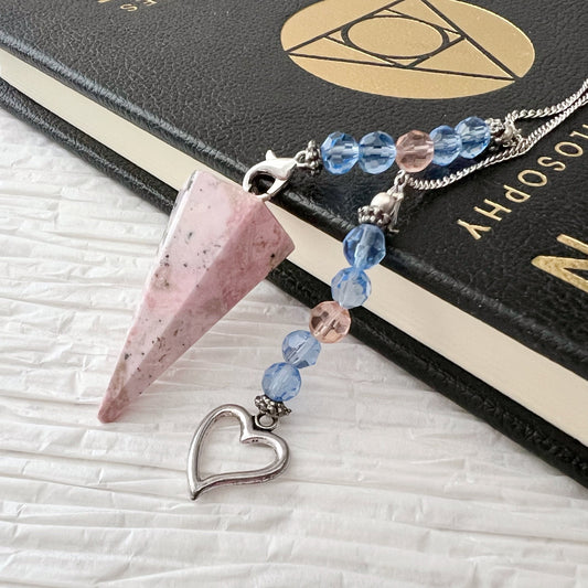 A Rhodonite Hexagonal Pendulum with Heart Charm lies atop a black book with gold text, embodying emotional healing. The chain includes a silver heart charm, adding elegance. The surface beneath the book and pendulum is textured white.