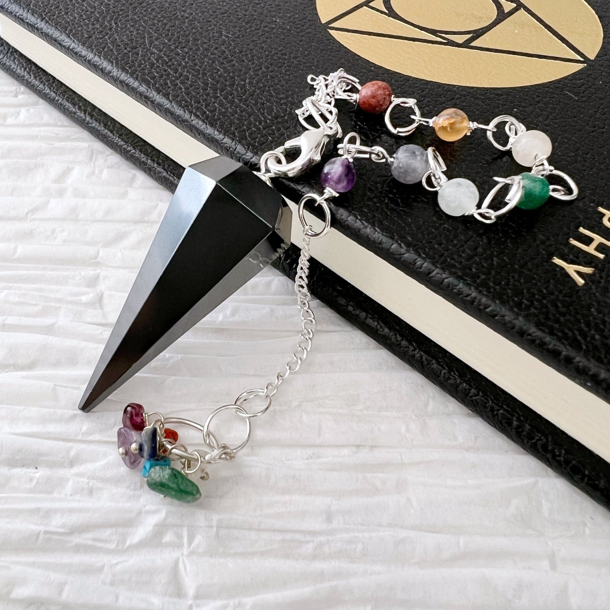 A Hematite Hexagonal Pendulum with a Chakra crystal chain rests on a black textured book with gold designs and text. The pendulum's point is placed at the edge of the book, and the colorful beads are arranged towards the bottom on a white surface.