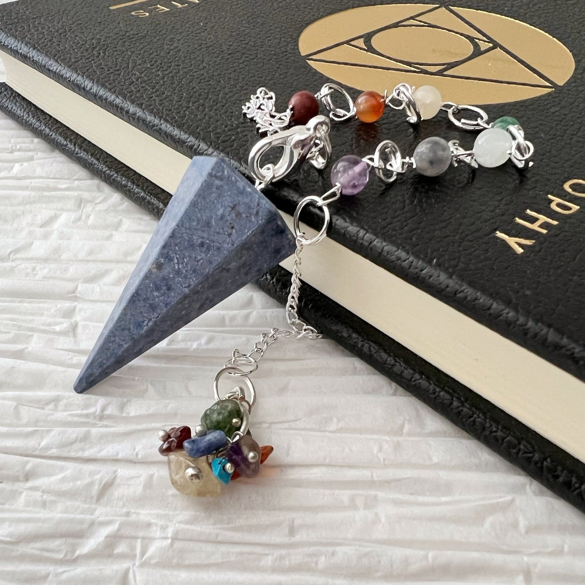 A Dumortierite Hexagonal Pendulum with a metal chain adorned with various colorful stones lies atop a hardcover book with a textured black cover. The book has a geometric design and text on the cover and rests on a white crinkled surface, adding to the aura of its healing properties and chakra crystals.