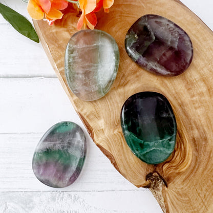 A wooden surface holds four smooth, oval-shaped Rainbow Fluorite Worry Stones in shades of green, purple, and clear. An orange flower rests at the top left corner, with green leaves extending from it, complementing the vibrant colors and aura cleansing properties of the stones.